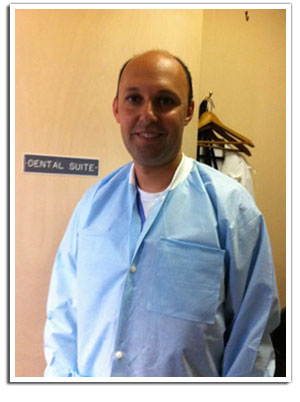 The image features a man wearing protective clothing, likely a medical gown, standing indoors with a sign for a dental suite visible behind him.