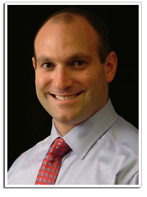 The image features a man with a bald head, wearing a dark shirt, smiling at the camera. He has short hair and appears to be indoors with a plain background.