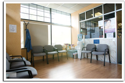 The image shows an interior view of a clean and well-lit medical waiting room with a window, chairs, and a desk.