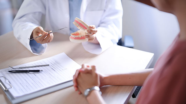 The image depicts a dental professional showing a patient s teeth with a magnifying glass during a consultation, with both individuals seated at a table with medical records and a clipboard.