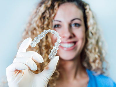 A woman wearing a blue shirt is holding up a transparent dental implant with her gloved hand against a white background, smiling at the camera.