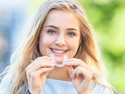 The image shows a young woman with blonde hair smiling at the camera while holding up a clear dental retainer between her teeth.