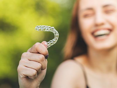 A young woman holding up a dental retainer with a smile on her face against a blurred outdoor background.