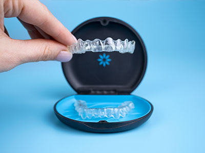 The image shows a person s hand holding an open dental impression tray containing a blue dental impression material with a visible tooth shape imprinted on it, placed against a blue background.