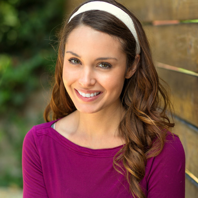 A young woman with long hair smiles at the camera, wearing a purple top and a white headband.