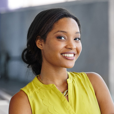 A young woman with dark hair smiling at the camera, wearing a yellow top with a light-colored blazer and a necklace, against a blurred background that suggests an urban setting.