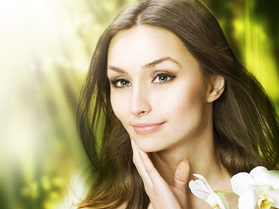 An image of a woman with light skin, long brown hair, and blue eyes. She has a neutral expression and appears to be wearing makeup. The background features soft lighting and blurred greenery, suggesting a natural setting.