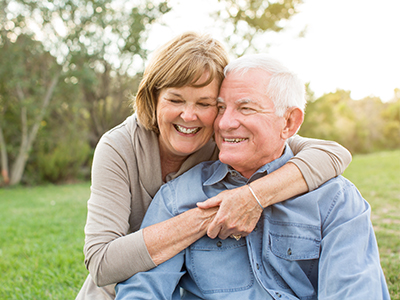 The image shows an elderly couple sharing a warm embrace, with the man seated and the woman standing behind him, both smiling and appearing content in each other s company.