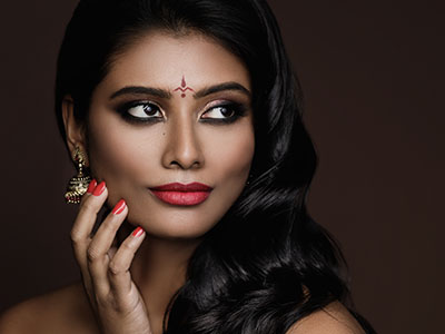 The image shows a woman with dark hair, wearing makeup including red lipstick and eye shadow, looking directly at the camera with her hand gently placed on her chin. She is wearing a sleeveless top and has a small bindi on her forehead.