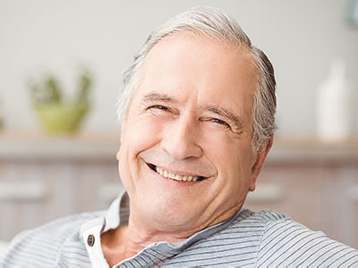 The image shows a man with gray hair, wearing glasses, smiling at the camera while seated comfortably indoors.